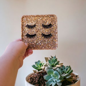 May include: A gold glitter tray with three pairs of false eyelashes. The tray is being held by a hand.