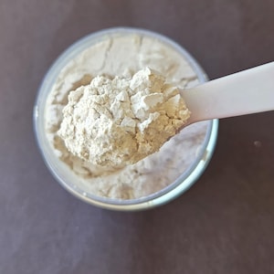 May include: A close-up shot of a clear container filled with a light beige powder. A white scoop holds a portion of the powder, showcasing its granular texture. The background is a solid, muted brown.
