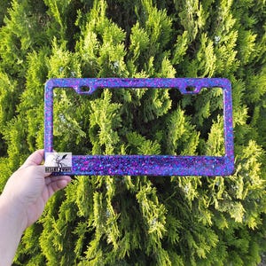 May include: A black license plate frame with a glittery finish in shades of purple and blue. The frame has a small logo in the bottom left corner that says "Desert Vinyl".
