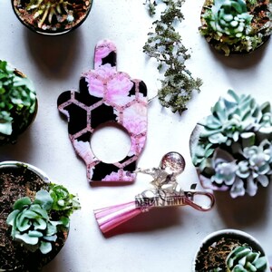 May include: A pink and black floral patterned keychain with a tassel and a small charm. The keychain is shaped like a middle finger.