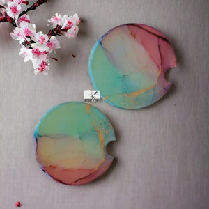 May include: Two round, colorful coasters with a cutout for a cup. The coasters have a blue, green, yellow, and pink marbled design with gold accents.