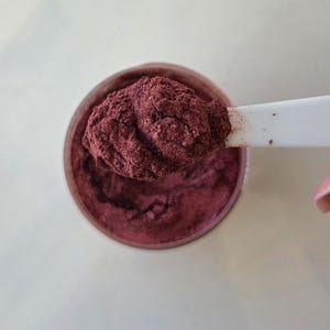 May include: A close-up shot of a jar filled with a deep burgundy powder. A white spoon holds a scoop of the same powder, showcasing its texture. The powder appears finely milled and has a slight shimmer.