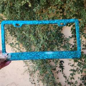 May include: A turquoise glitter license plate frame. The frame is made of metal and has a shiny, sparkly finish.