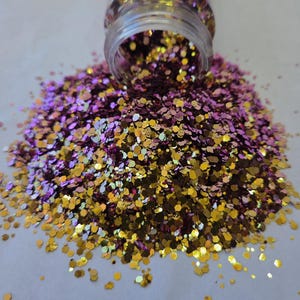 May include: A clear jar tipped over, spilling a cascade of glitter. The glitter is a mix of gold, purple, and iridescent colors, creating a shimmering effect. The glitter is in a hexagonal shape. Ideal for crafts and decorations.