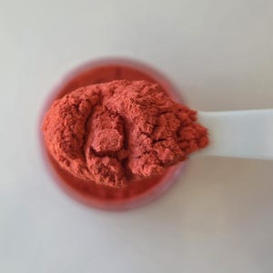 May include: A close-up shot of a white spoon holding a scoop of vibrant, reddish-orange powder. The powder appears finely ground and textured. The spoon is positioned over a small, clear container filled with the same powder.