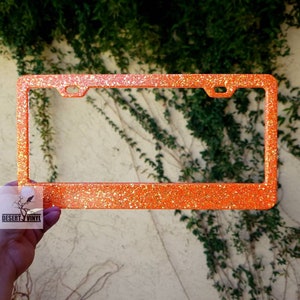 May include: Orange glitter license plate frame. The rectangular frame has a sparkly, glittery texture. The frame has pre-drilled holes for mounting. The logo "DESERT VINYL" is visible in the lower left corner.