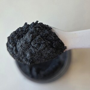 May include: Close-up of a spoonful of dark gray powder, likely a cosmetic pigment or dye. The powder has a textured, granular appearance and is held in a white plastic spoon. A dark container is visible below the spoon.