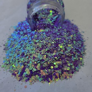 May include: A pile of iridescent glitter spills from a clear jar. The glitter is a mix of purple, blue, green, and gold, with a hexagonal shape. The glitter is scattered on a light gray surface.