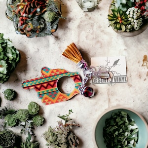 May include: A colorful, geometric hand-shaped keychain with a key ring and a tassel. The keychain is made of a hard plastic material and has a bright, vibrant design. The text "DESERT VINYL" is printed on a small card attached to the keychain.