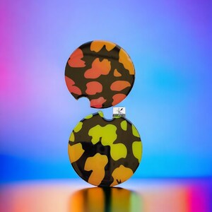 May include: Two round coasters with a black background and a colorful abstract pattern. The top coaster is orange and pink, and the bottom coaster is yellow and green.