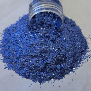 May include: A close-up of a pile of blue glitter spilling from a clear jar. The glitter is a deep blue color with iridescent flecks, creating a sparkling effect. The glitter is in a hexagonal shape.