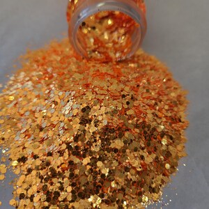 May include: A clear plastic jar tipped over, spilling a pile of orange and gold glitter. The glitter is in various shapes and sizes, reflecting light. The glitter is scattered on a light gray surface.