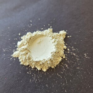 May include: A small pile of light green powder, possibly a cosmetic product or pigment, sits on a dark gray surface. The powder is finely ground, with a slightly textured appearance. Some powder has scattered around the main pile.