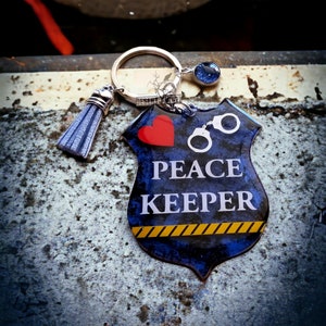 May include: A blue and silver keychain with a peace sign and the words "Peace Keeper". The keychain also features a red heart and handcuffs.