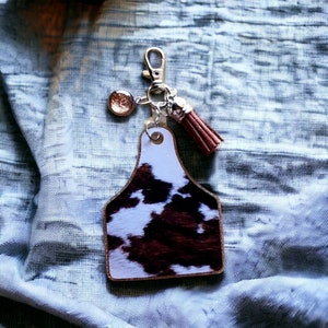 May include: Cowhide keychain with a brown tassel and a glittery charm. The keychain is shaped like a cattle ear tag.