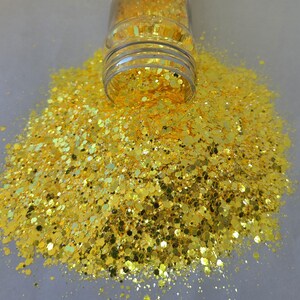 May include: A clear plastic jar tipped over, spilling a large pile of yellow glitter. The glitter is a mix of shapes and sizes, reflecting light. The glitter is scattered on a white surface.