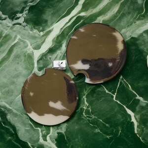 May include: Two round, brown and white coasters with a cut-out design. The coasters are on a green marble surface.