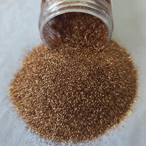May include: A clear plastic jar tipped over, spilling a pile of bronze glitter onto a white surface. The glitter is finely ground and reflects light, creating a sparkling effect. Ideal for crafts and decorations.