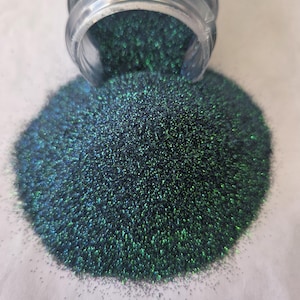 May include: A clear jar with iridescent glitter spilling out onto a white surface. The glitter is a mix of dark blue and green, creating a shimmering effect. The glitter is fine and appears to be cosmetic grade.