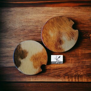 May include: Two brown and beige round coasters with a cutout on the side. The coasters are on a wooden surface. The text "DESERT VINYL" is on a white label with a black silhouette of a bird on a branch.