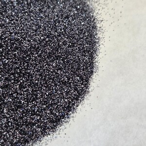 Puede incluir: Primer plano de un mont&oacute;n de purpurina gris oscuro con una superficie brillante y reflectante. La purpurina est&aacute; densamente empaquetada, con part&iacute;culas individuales que captan la luz. La purpurina est&aacute; sobre una superficie blanca.