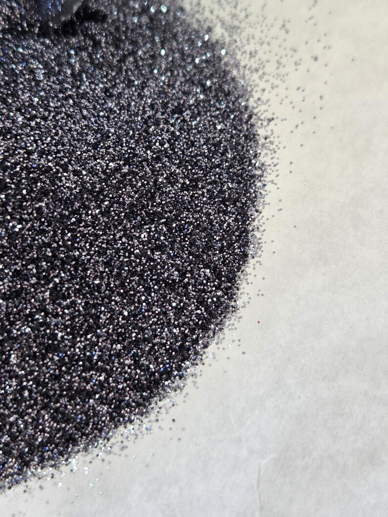 Puede incluir: Primer plano de un mont&oacute;n de purpurina gris oscuro con una superficie brillante y reflectante. La purpurina est&aacute; densamente empaquetada, con part&iacute;culas individuales que captan la luz. La purpurina est&aacute; sobre una superficie blanca.