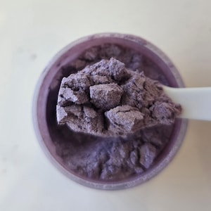 May include: Close-up of a container filled with a shimmering, lavender-colored powder. A white spoon holds a portion of the powder, showcasing its textured, granular appearance. The container is a matching lavender color, creating a cohesive look.