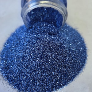 May include: A clear glass jar tipped over, spilling a large pile of sparkling blue glitter. The glitter is a deep, rich blue with a shimmering, reflective quality. The glitter is scattered on a white surface.