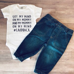May include: A white baby bodysuit with the text "GOT MY MIND ON MY MOMMY AND MY MOMMY ON MY MIND #LAIDBACK" alongside a pair of blue denim baby trousers. The trousers have an elasticated waistband and cuffs.