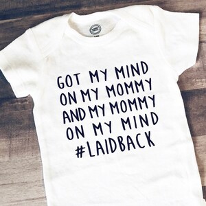 May include: White baby bodysuit with dark blue text reading "GOT MY MIND ON MY MOMMY AND MY MOMMY ON MY MIND #LAIDBACK". The garment has short sleeves and a round neck. The Wonder Nation logo is visible.