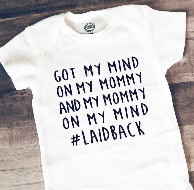 May include: White baby bodysuit with dark blue text reading "GOT MY MIND ON MY MOMMY AND MY MOMMY ON MY MIND #LAIDBACK". The garment has short sleeves and a round neck. The Wonder Nation logo is visible.