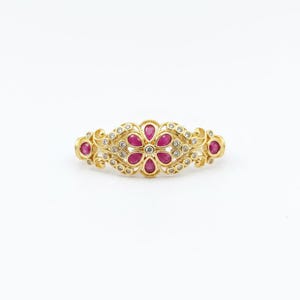 May include: A gold ring featuring a central floral motif composed of six pear-shaped red rubies surrounding a small clear stone. The band is decorated with intricate gold scrollwork and accented with numerous small, round, clear white stones.