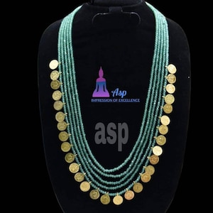 Puede incluir: Un collar de múltiples hilos con cuentas verdes y detalles de monedas de oro. El collar está en un busto de maniquí negro. El texto "asp Impression of Excellence" es visible en el busto.