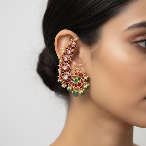May include: An ear cuff earring featuring a gold-tone metal frame adorned with teardrop-shaped red stones, circular green stones, and small white pearls. The design follows the curve of the ear, with dangling elements at the base of the piece.