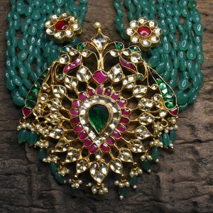May include: A gold necklace with a large, ornate pendant featuring green and pink gemstones. The pendant is surrounded by a string of green beads.