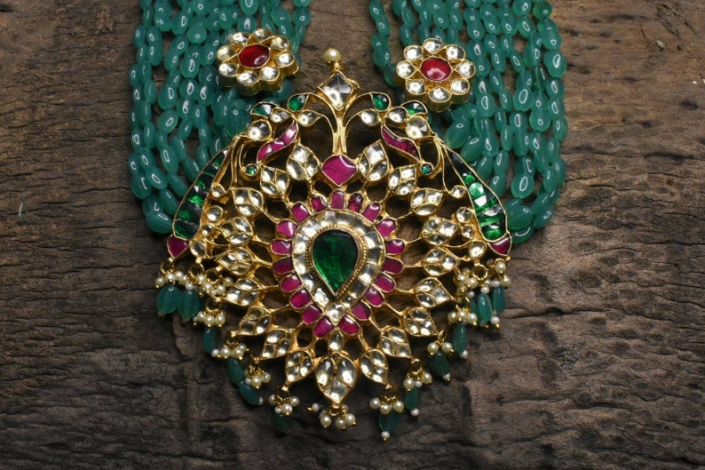 May include: A gold necklace with a large, ornate pendant featuring green and pink gemstones. The pendant is surrounded by a string of green beads.