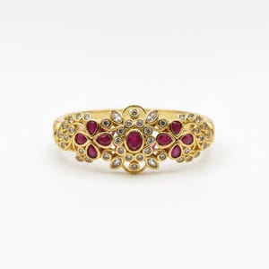 May include: A gold ring featuring a central oval-cut ruby surrounded by small round white stones. Two floral-shaped clusters, each containing four pear-cut rubies, flank the center. The entire band is decorated with intricate gold filigree details.