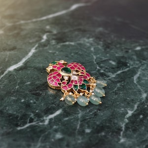 May include: A gold-toned brooch featuring a stylized floral design with inlaid pink and green stones. The piece is accented with small white beads and finished with five dangling, pale blue-green oval beads suspended from the bottom edge.