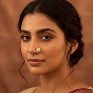 May include: A gold-toned nose ring featuring a semi-circular design adorned with small white pearls and clear stones. The jewelry piece is positioned on the side of the nostril, showcasing a delicate metal frame with a cluster of pearls at the center.
