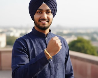 Può includere: Una persona indossa un turbante blu navy e un kurta abbinato. Un braccialetto color oro è visibile al polso. Lo sfondo è sfocato, con un paesaggio urbano in lontananza.