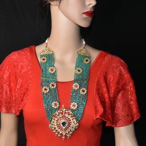 May include: A long, multi-strand necklace with green beads and red and gold accents. The necklace has a large, ornate pendant with a green stone in the center.
