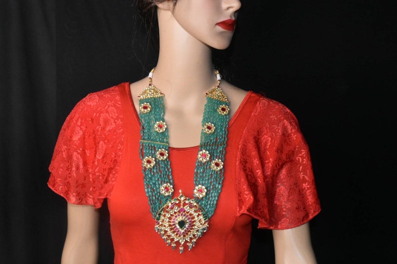 May include: A long, multi-strand necklace with green beads and red and gold accents. The necklace has a large, ornate pendant with a green stone in the center.