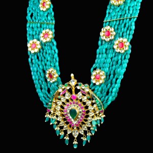 May include: A turquoise and gold necklace with a large, ornate pendant featuring a floral design and colorful gemstones. The pendant is surrounded by strands of turquoise beads.