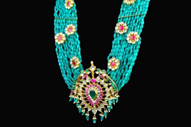 May include: A turquoise and gold necklace with a large, ornate pendant featuring a floral design and colorful gemstones. The pendant is surrounded by strands of turquoise beads.