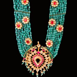May include: A multi-strand necklace made of green beads with a large, ornate pendant featuring red and white gemstones. The pendant is in the shape of a stylized flower.