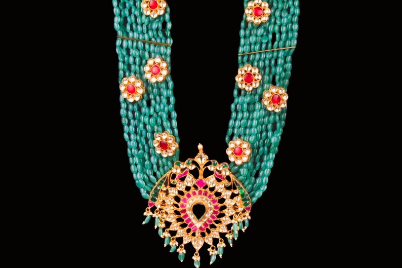 May include: A multi-strand necklace made of green beads with a large, ornate pendant featuring red and white gemstones. The pendant is in the shape of a stylized flower.