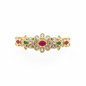 May include: A gold ring featuring a central oval red gemstone surrounded by clear white stones in a floral arrangement. The design includes additional small green and red stones set within the gold band, creating a symmetrical pattern.
