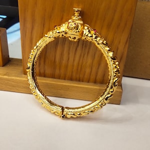 May include: A gold-toned hinged bangle bracelet featuring an ornate, textured surface with intricate scrollwork patterns. The top section includes a decorative centerpiece adorned with small, embedded clear and red stones, providing a metallic finish.