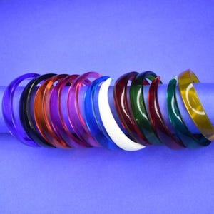 May include: A collection of colorful, translucent bangles in various shades, including purple, black, orange, pink, blue, white, red, green, and yellow. The bangles are arranged on a purple surface, showcasing their vibrant hues and glossy finish.
