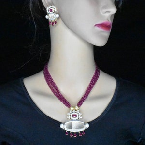 Puede incluir: Un collar rosa y blanco con un colgante que presenta un gran rubí y diamantes más pequeños. El collar está hecho de múltiples hebras de pequeñas cuentas rosas. El colgante tiene la forma de un rectángulo con la parte superior e inferior redondeadas. El colgante está rodeado por un borde de pequeños diamantes. El collar lo lleva un maniquí con una camisa negra.